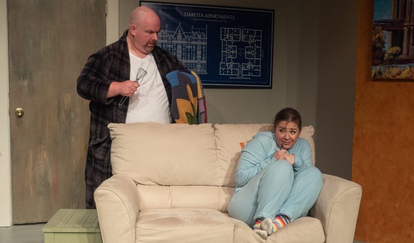 One actor sitting frightened on a couch, while the other confronts her with a whisk.