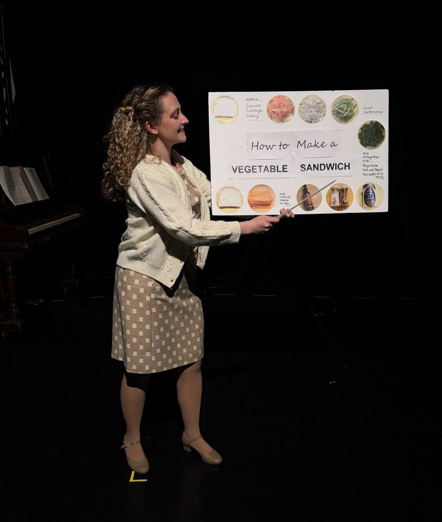 A woman in a beige sweater and tan polka dot dress holds a sign and points to instructions on how to make a sandwich.