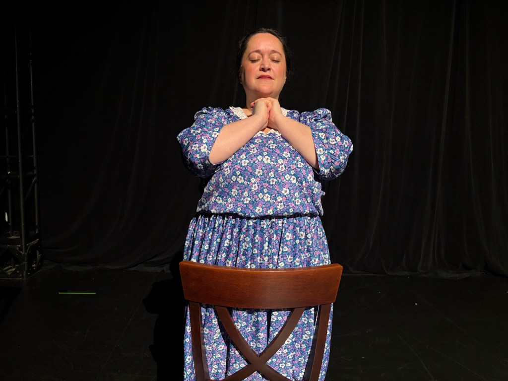 A woman wearing a blue flowered dress prays in front of a chair.